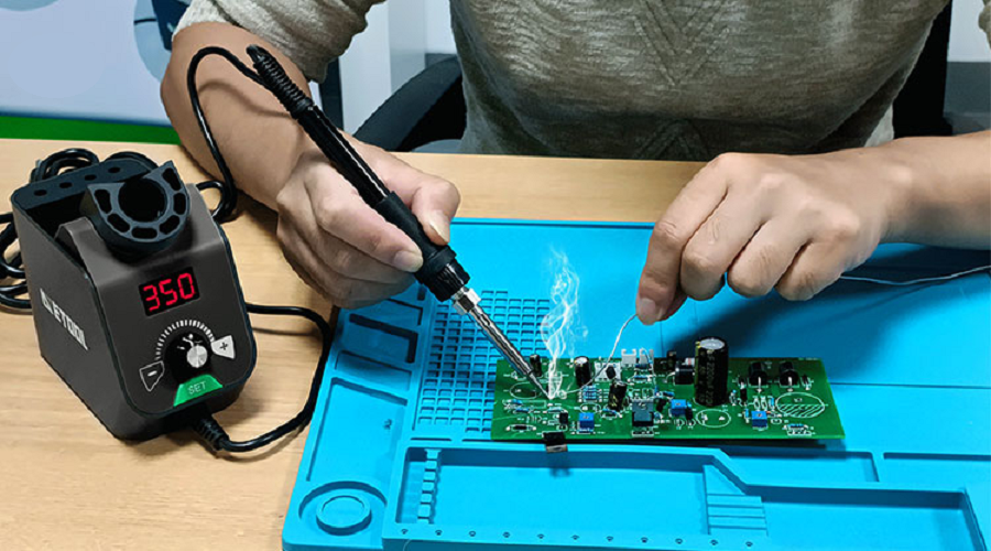 Person using an electric soldering iron to repair a green circuit board on a blue mat