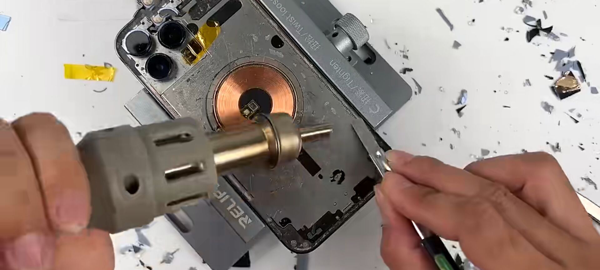 Hands using heat gun and tweezer to repair smartphone wireless charging coil on workbench