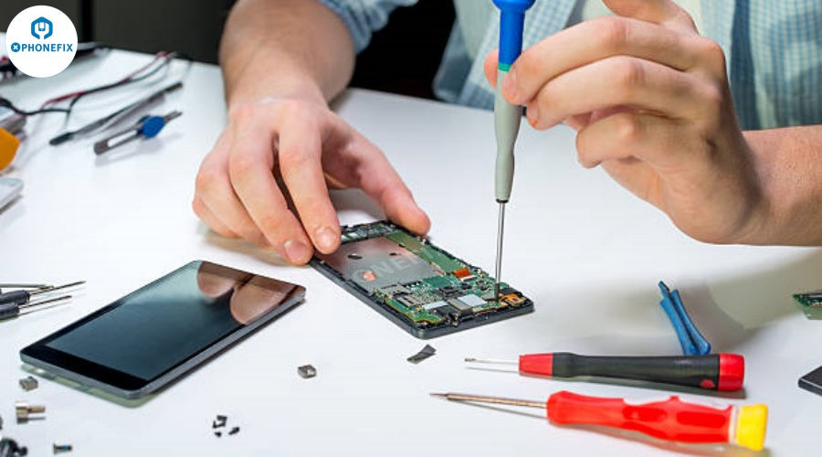 Technician using screwdriver to repair smartphone motherboard with phone repair tools on table.